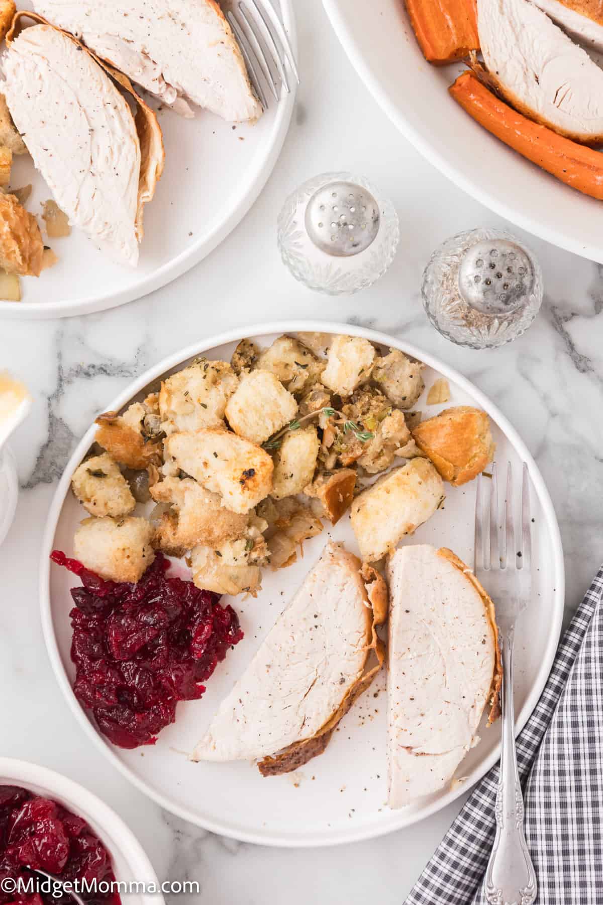 A plate with sliced turkey, stuffing, and cranberry sauce. Two salt and pepper shakers are nearby on a white marble surface.