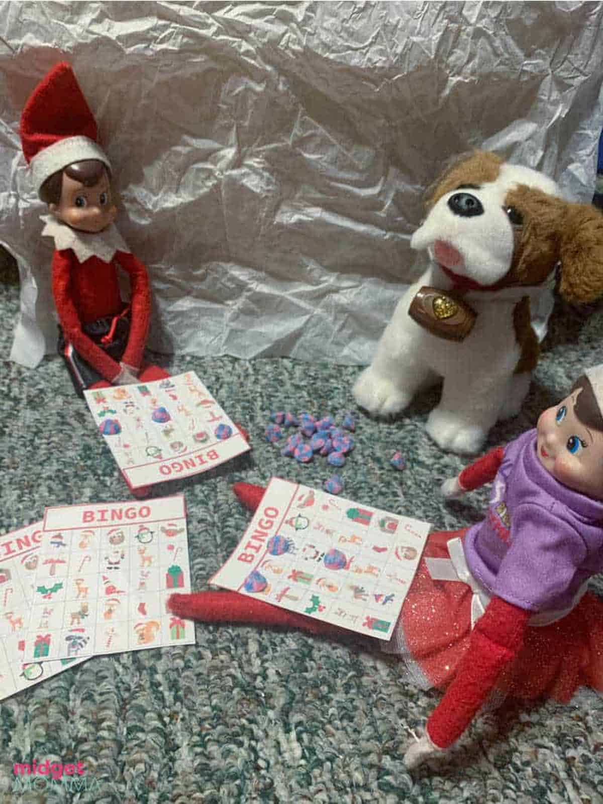 An elf on the shelf with two dolls and a dog.