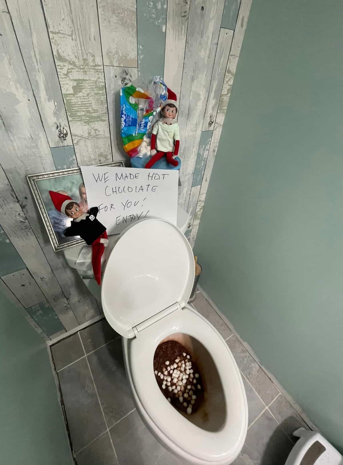 An elf on the toilet with a sign on it.