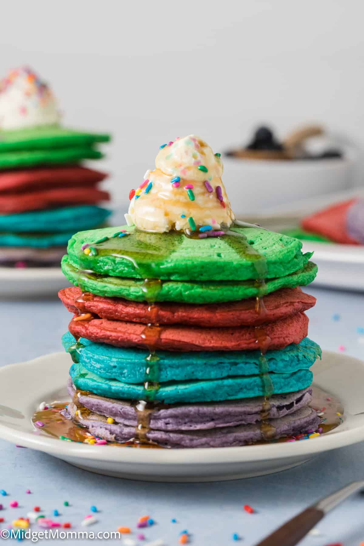 Stack of Rainbow pancakes with maple syrup, whipped cream and sprinkles on a plate.