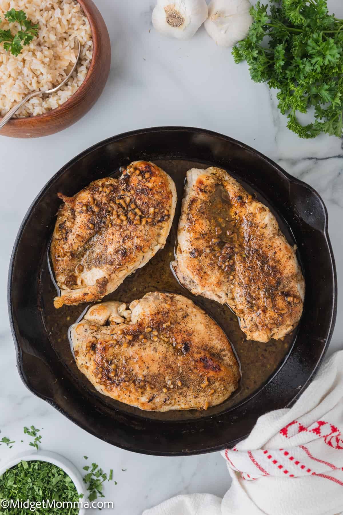 A cast iron skillet containing three seasoned and cooked pork chops, surrounded by a bowl of rice, parsley, garlic bulbs, and a cloth on a marble surface.