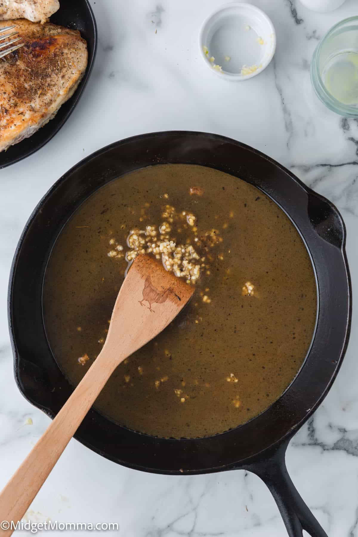 A wooden spoon stirring a dark brown sauce with minced garlic in a black skillet. A plate with cooked chicken breasts is partially visible in the background.