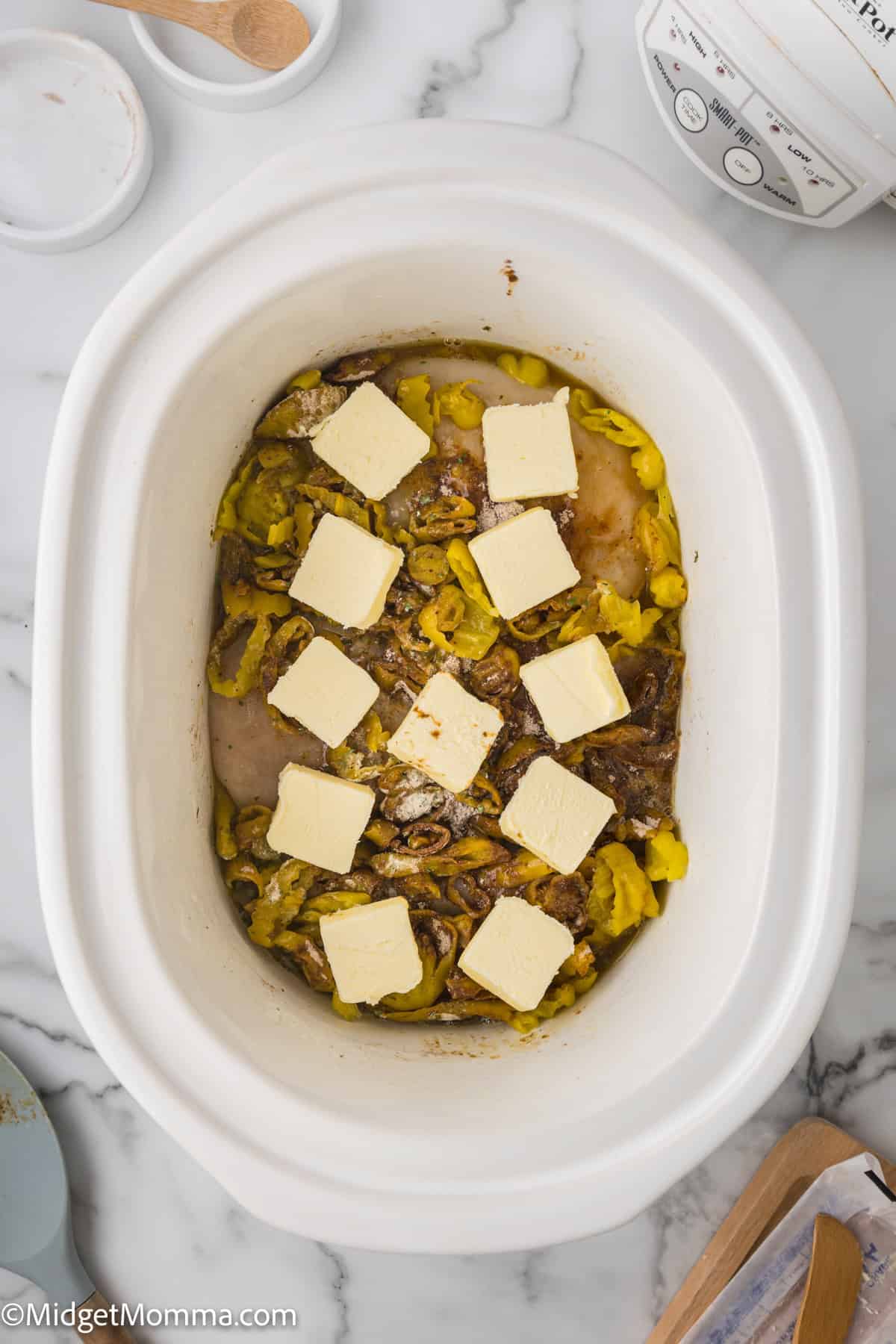 A white slower cooker with raw chicken, seasoning, peppers and butter