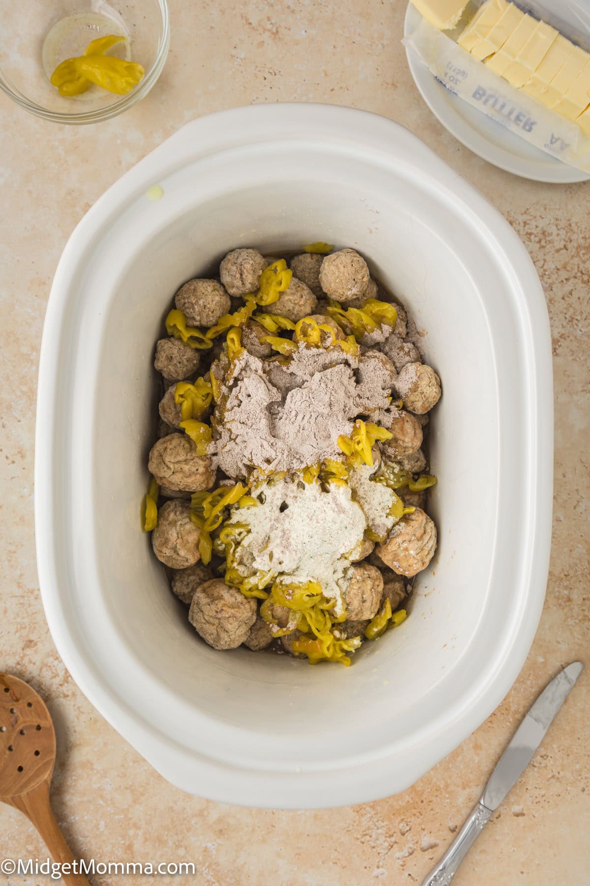 A slow cooker filled with raw meatballs, seasoning, and peppers. A wooden spoon, butter, and butter wrapper are placed next to it.