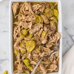 A rectangular dish filled with shredded mississippi pork roast, accompanied by a serving spoon. A small bit of napkin and part of a mashed potato dish are visible at the edges.