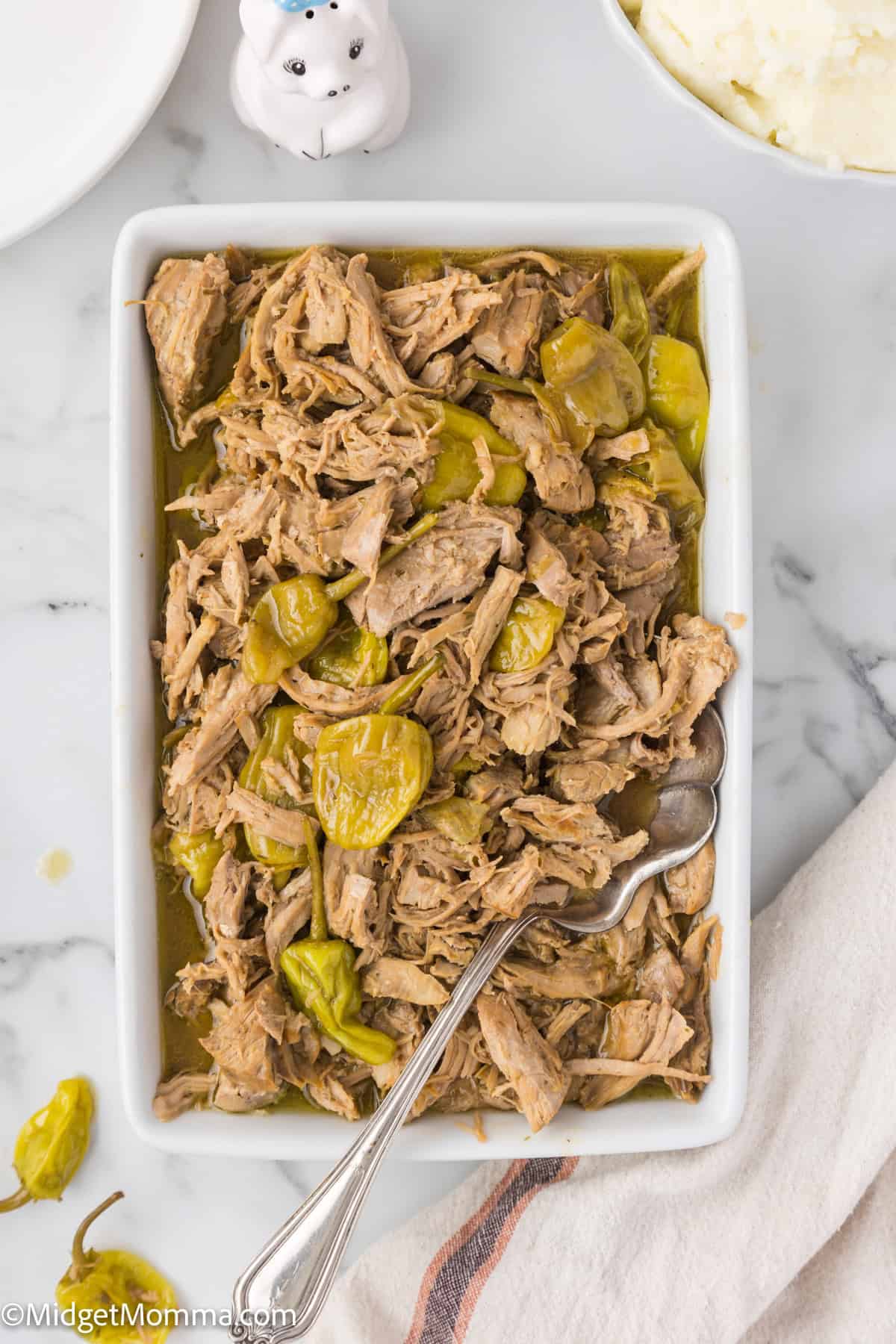 A rectangular dish filled with shredded mississippi pork roast, accompanied by a serving spoon. A small bit of napkin and part of a mashed potato dish are visible at the edges.