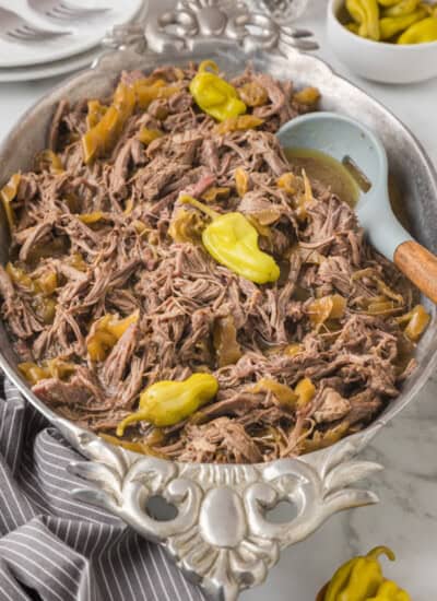 A serving dish filled with shredded beef, green peppers, and a bit of broth. A serving spoon is placed on the side. Plates, cutlery, and a bowl of additional green peppers are in the background.