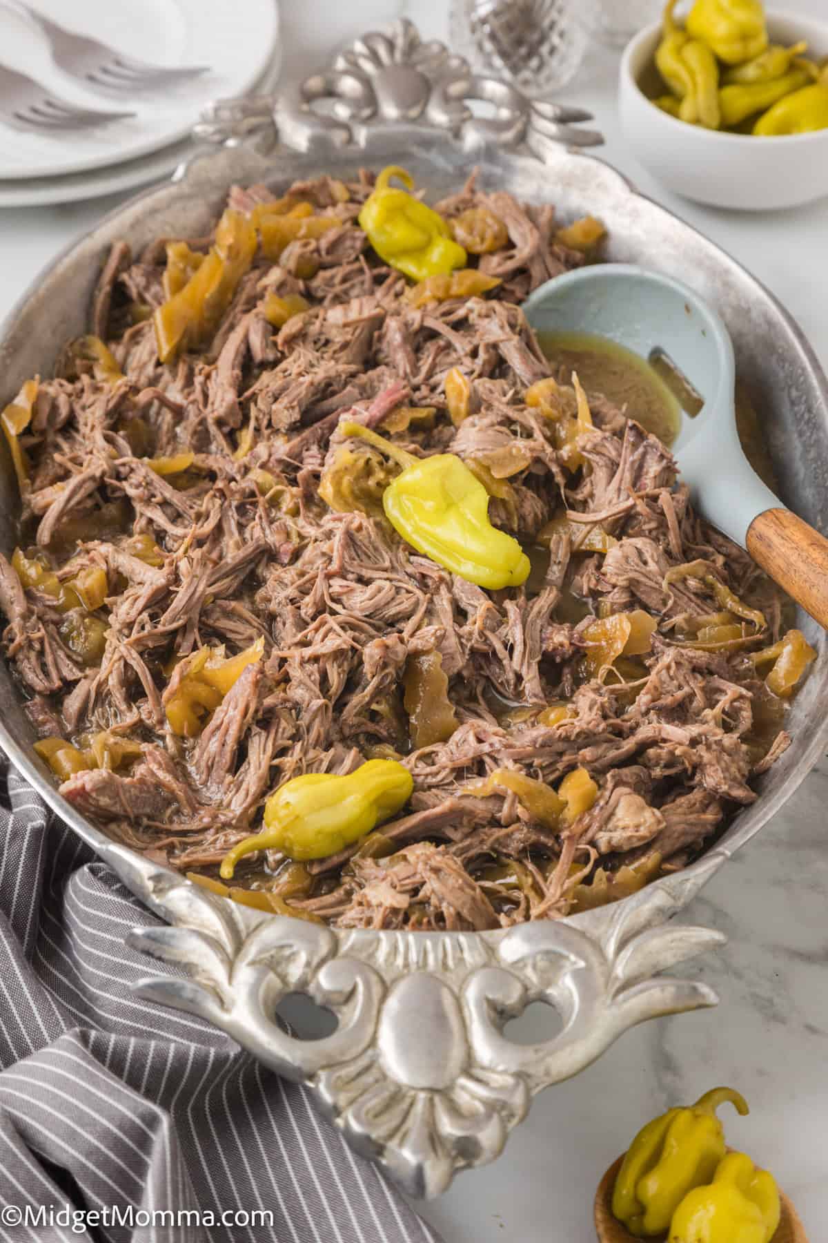 A serving dish filled with shredded beef, green peppers, and a bit of broth. A serving spoon is placed on the side. Plates, cutlery, and a bowl of additional green peppers are in the background.