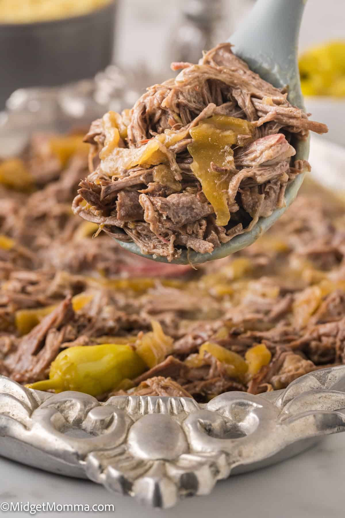 A spoon holding a portion of shredded beef with peppers over a plate filled with the same dish. An engraved silver plate holds the beef.