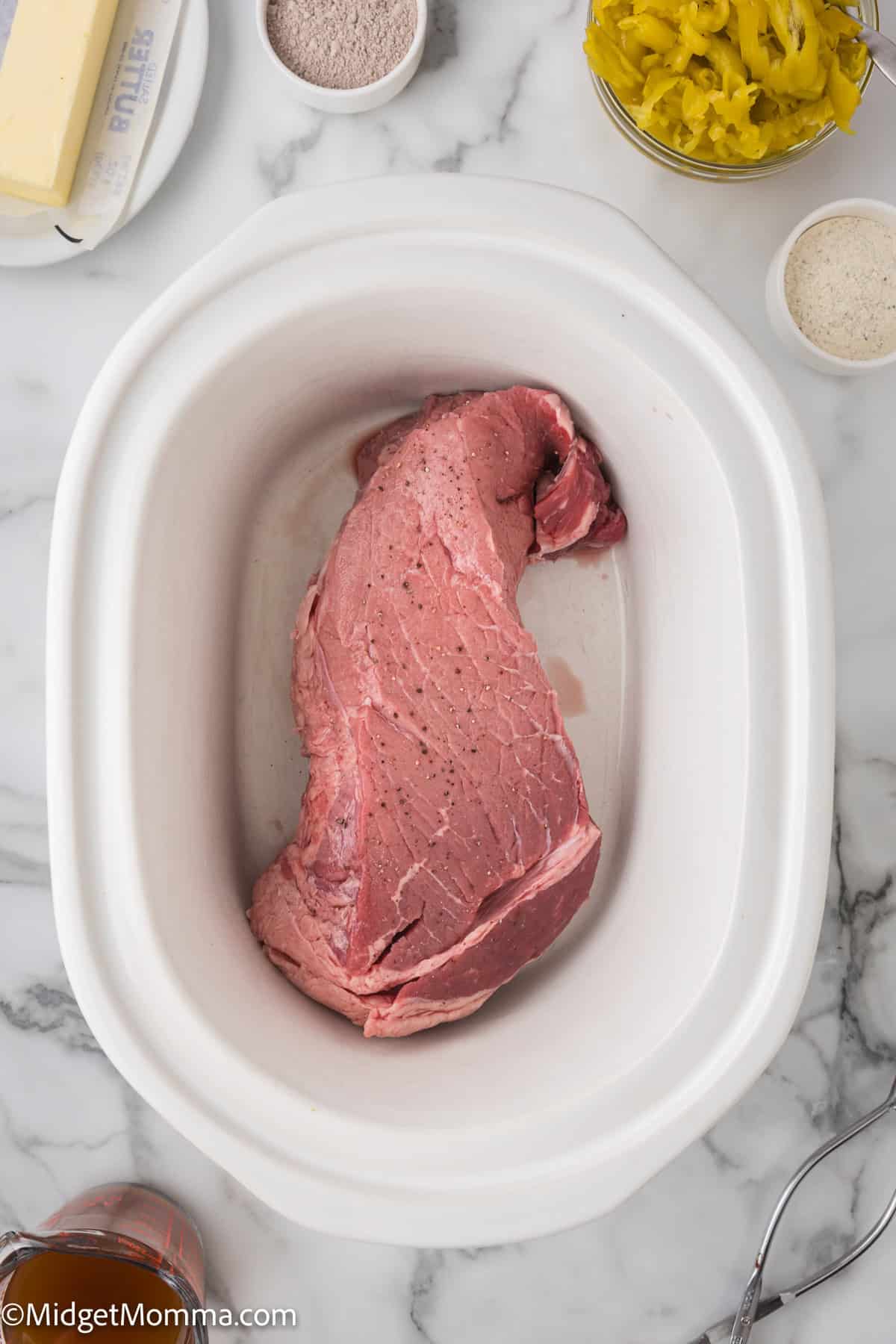 Raw beef placed inside a white slow cooker, surrounded by various ingredients including butter, sliced peppers, and seasoning in small bowls on a marble countertop.