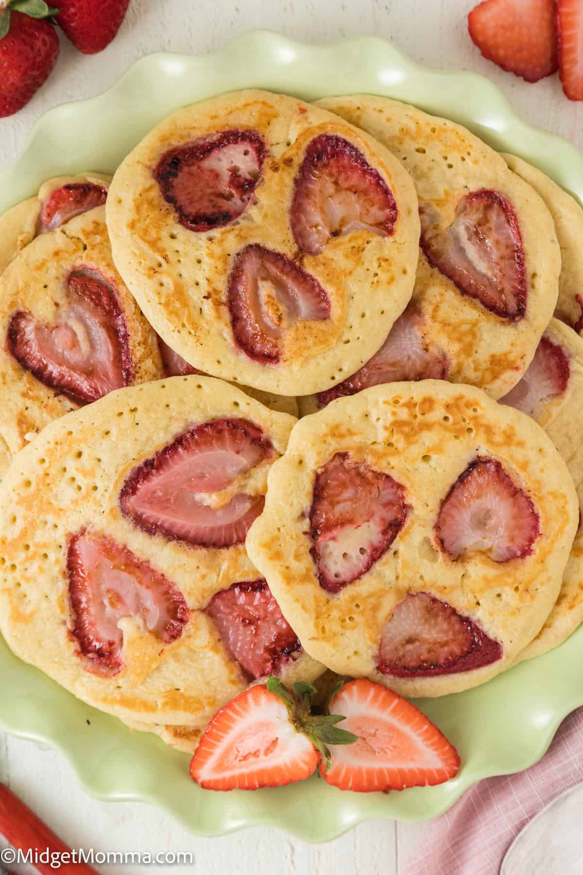 A plate of fluffy pancakes with sliced strawberries embedded in them, surrounded by fresh strawberry halves on a light-colored surface.