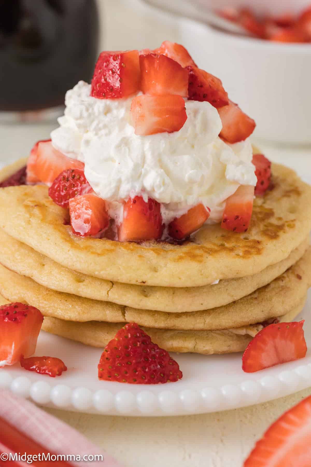 A stack of pancakes topped with whipped cream and fresh strawberry pieces on a white plate. Additional strawberry pieces are scattered around the plate.