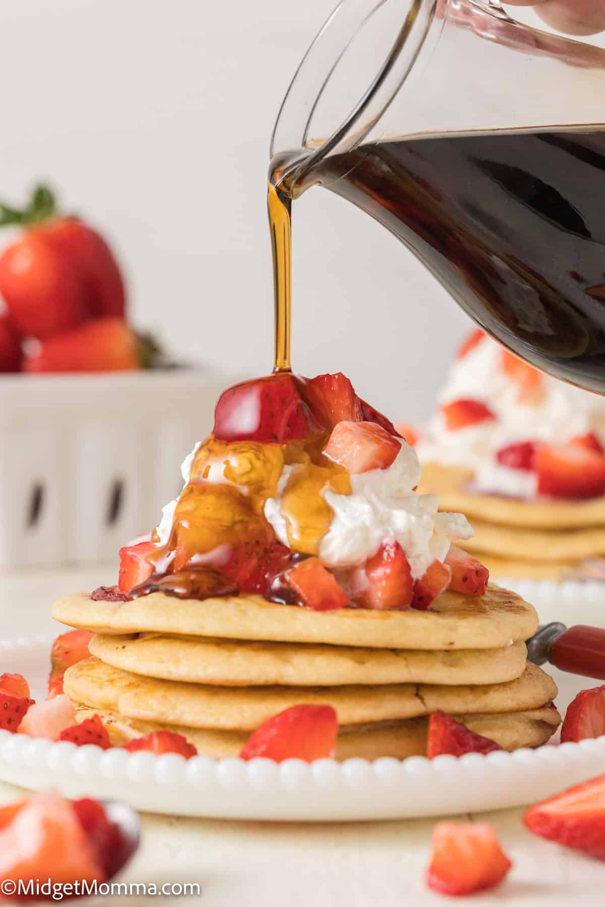 A stack of pancakes topped with whipped cream and chopped strawberries is being drizzled with syrup from a glass pitcher.