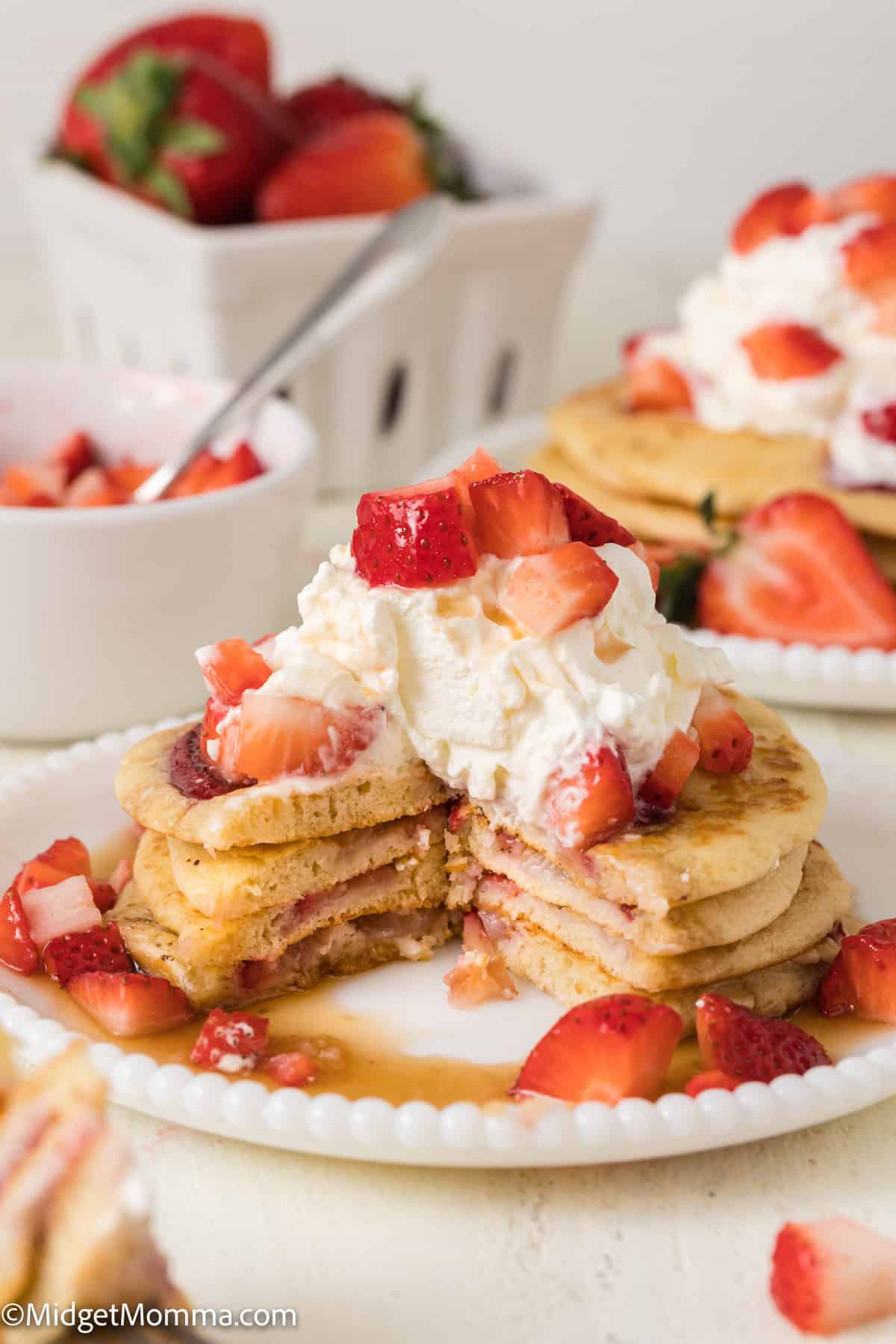 A stack of pancakes with whipped cream and chopped strawberries on top, partially sliced to display layers, accompanied by a white dish of whole strawberries in the background.