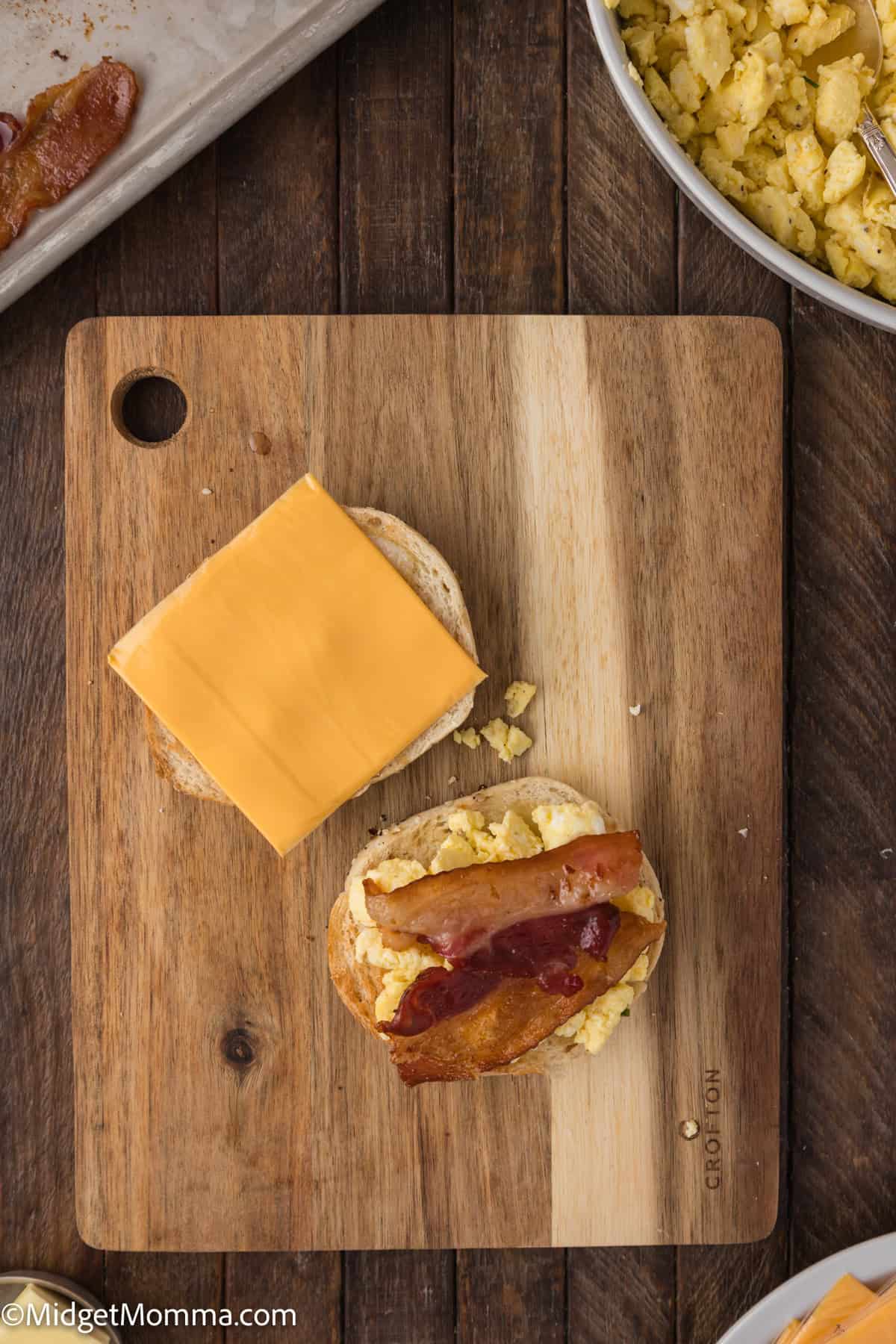A sandwich being prepared on a wooden cutting board, consisting of scrambled eggs, bacon, a slice of cheese, and jam on a bun. Additional scrambled eggs are visible in a bowl nearby.