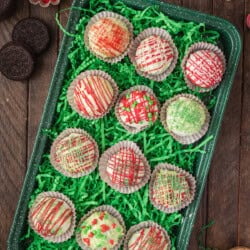 A tray of decorative holiday-themed cake balls with drizzled icing, placed on green shredded paper, surrounded by small bowls of colored sugar and festive sprinkles.
