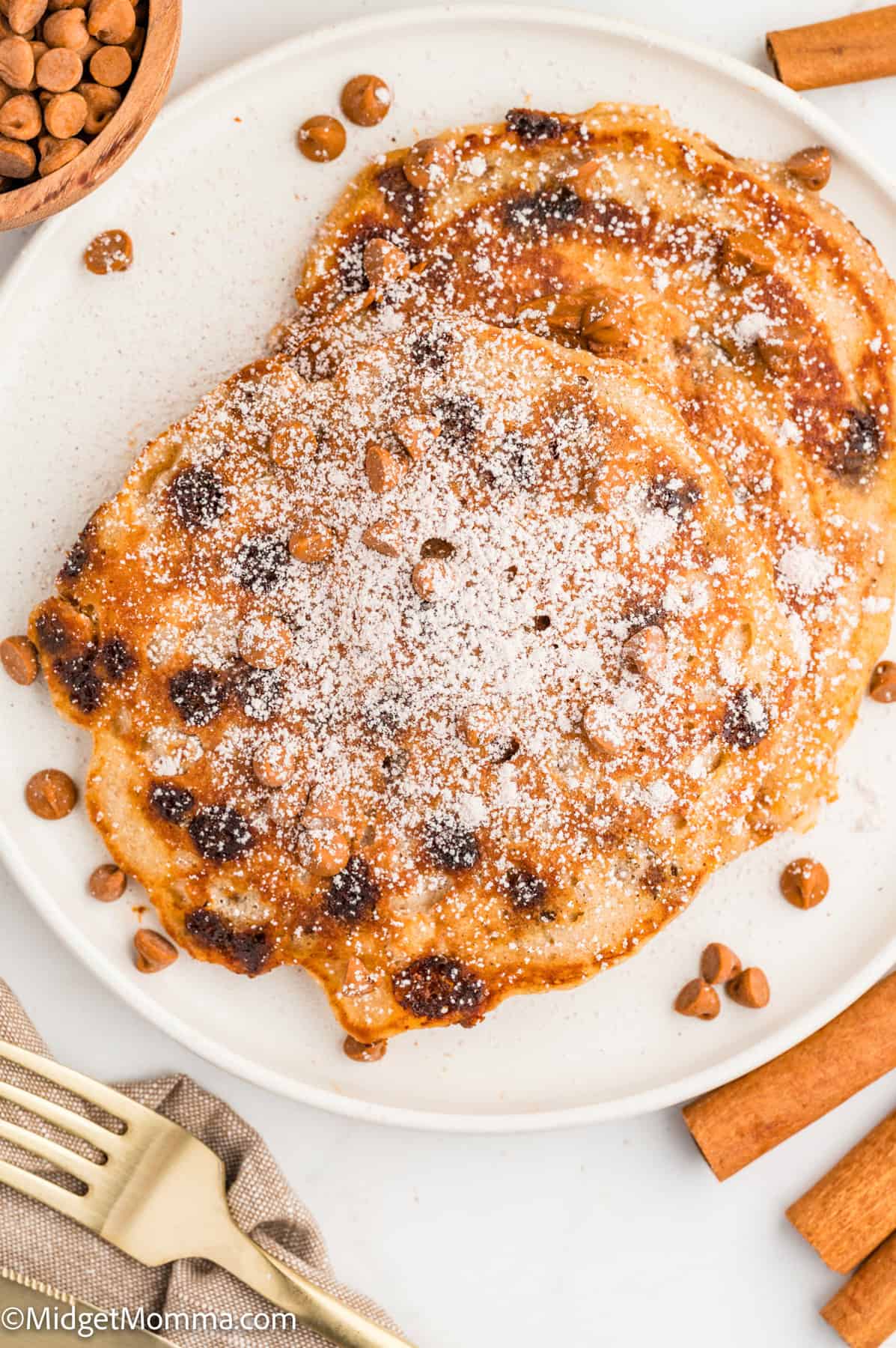 Two pancakes dusted with powdered sugar on a white plate, surrounded by cinnamon sticks and scattered chocolate chips.