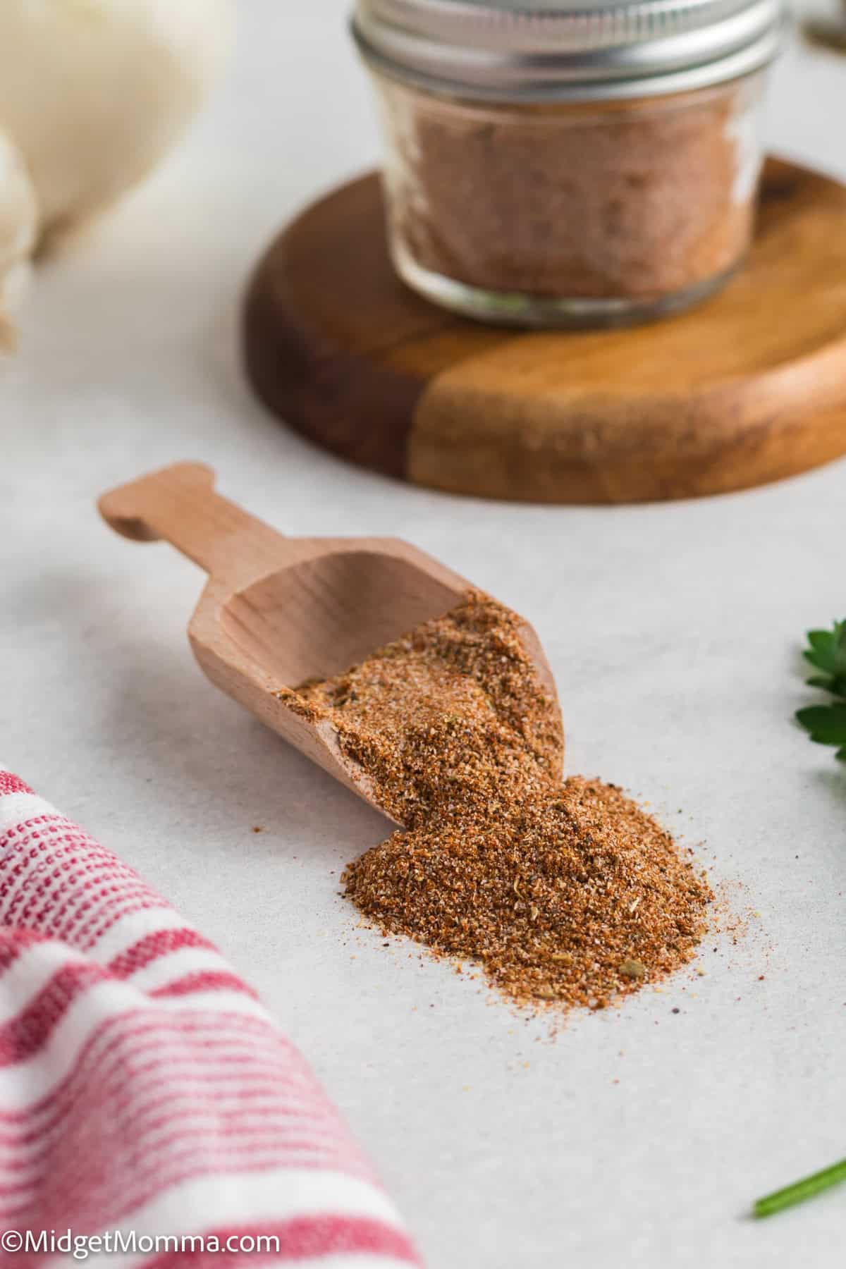 A wooden scoop with adobo seasoning spilled on a countertop. A jar of seasoning is in the background, along with a red-striped cloth and parsley.