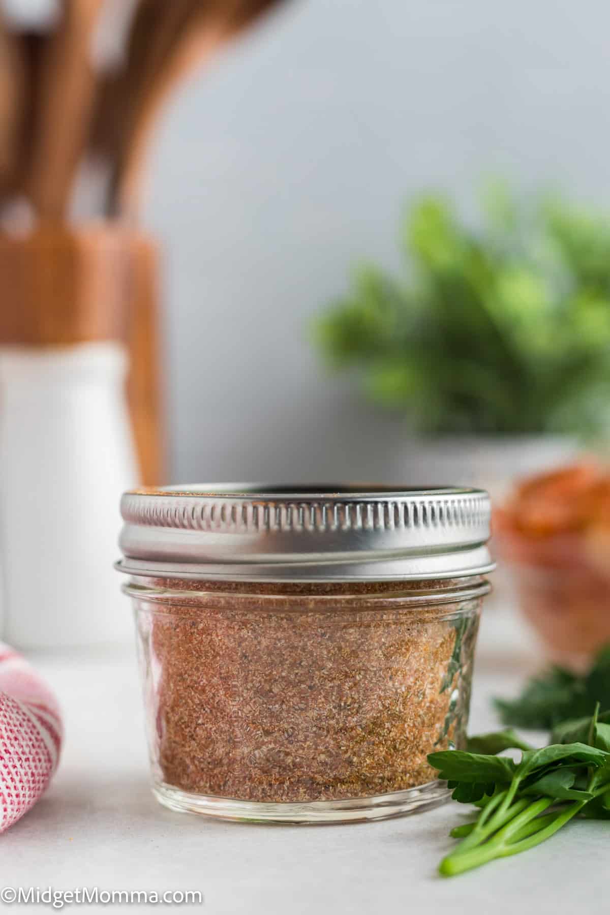 A small glass jar filled with adobo seasoning mix, sealed with a metal lid, sits on a kitchen counter. Fresh herbs are blurred in the background.