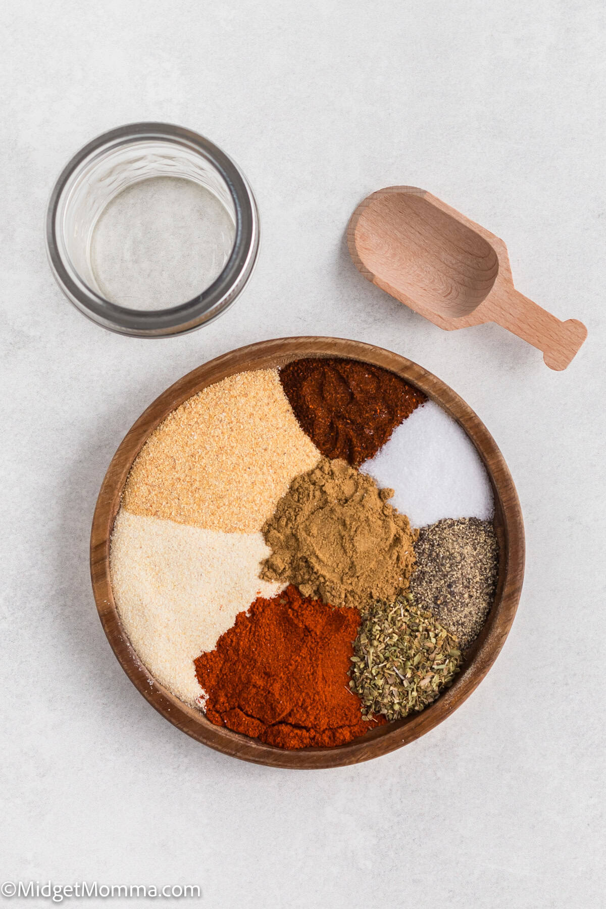 Adobo Seasoning Recipe ingredients. A wooden bowl with various spices arranged in sections, a small wooden scoop, and an empty jar nearby on a light surface.