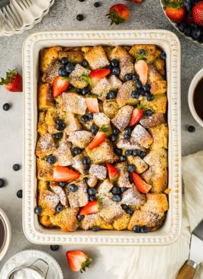 Baked French toast casserole topped with powdered sugar, strawberries, and blueberries in a rectangular dish, surrounded by fresh fruit and a cup of coffee.