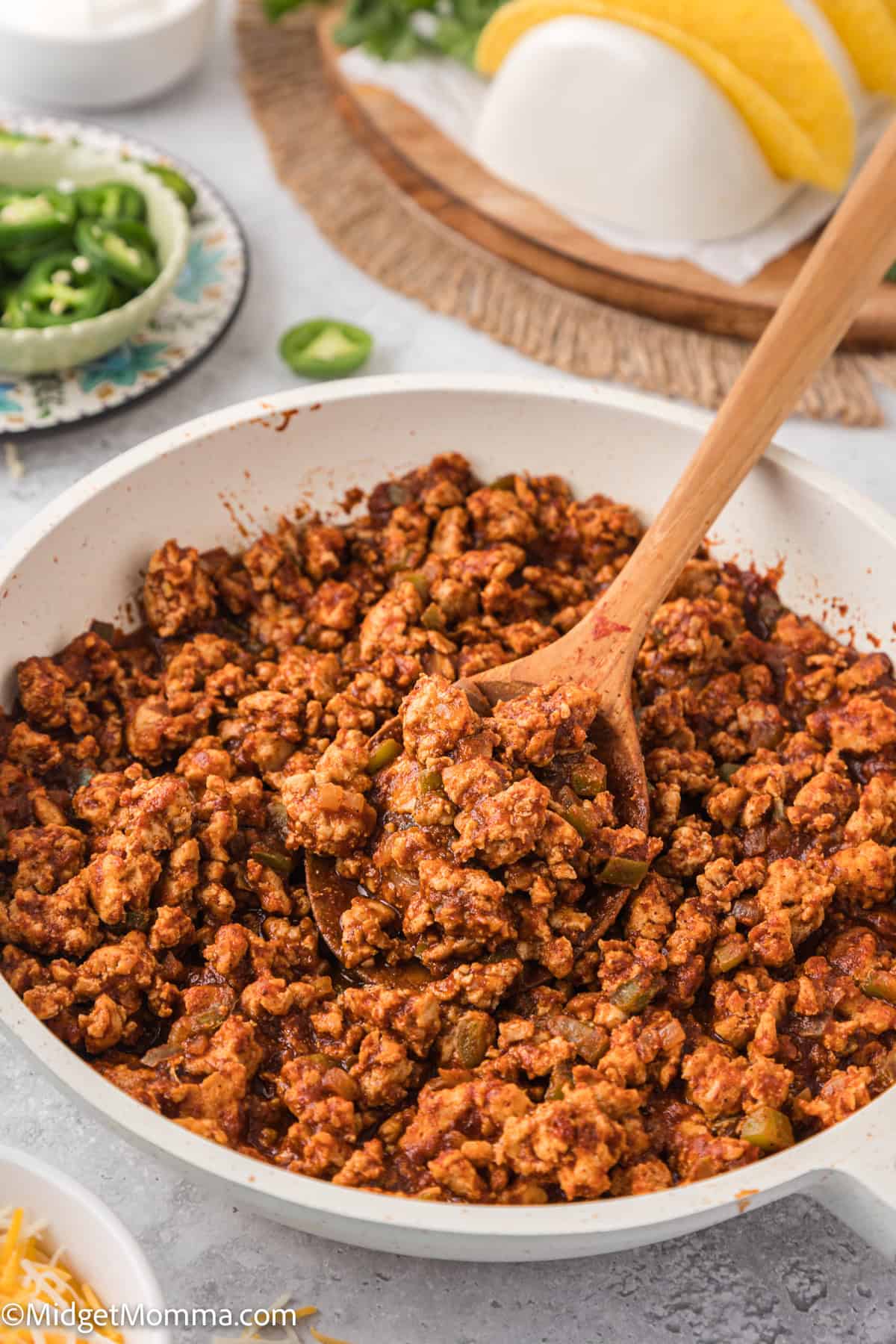 A skillet with seasoned ground meat and chopped vegetables, stirred with a wooden spoon. Taco shells and sliced jalapeños are visible in the background.