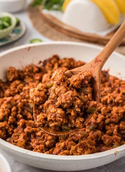 A wooden spoon holds a portion of seasoned ground meat from a white pan. Tacos and greens are blurred in the background.