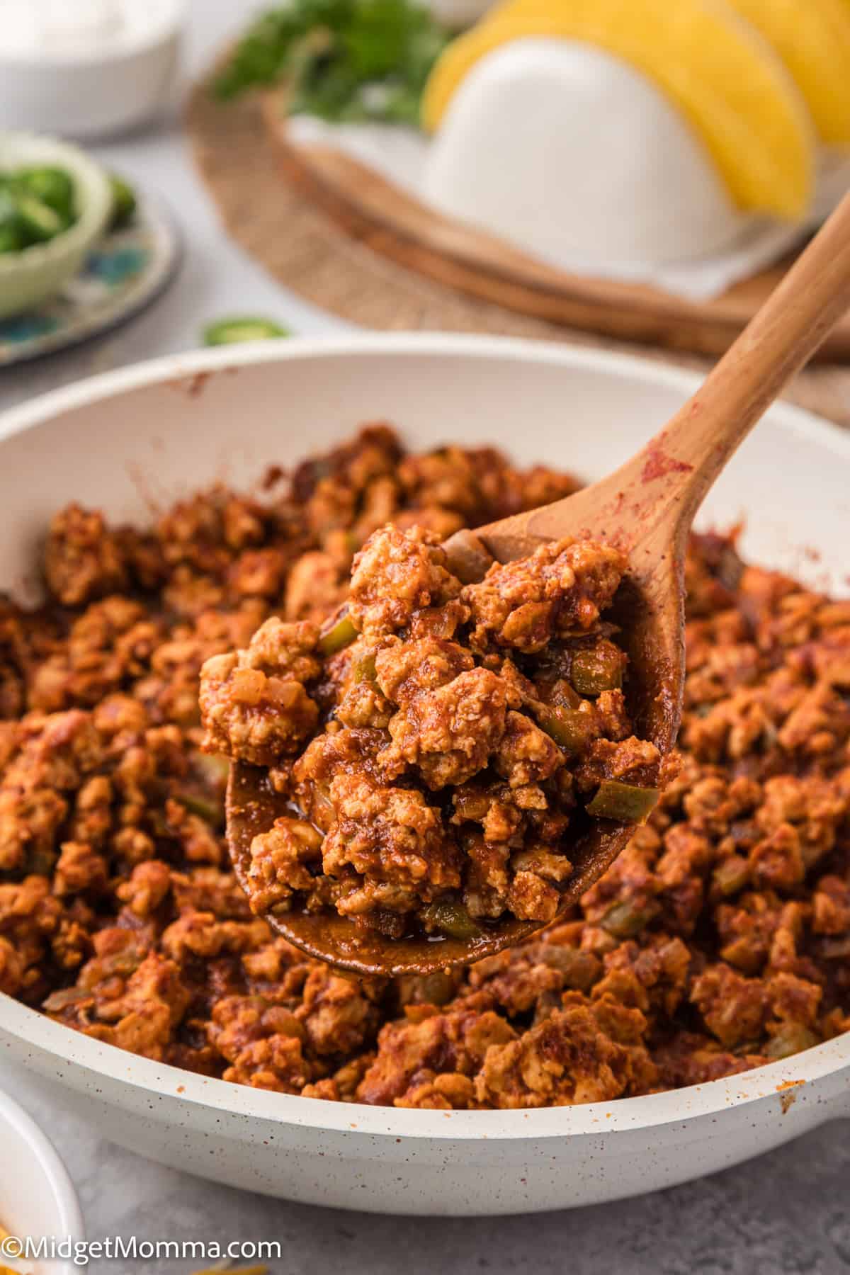 A wooden spoon holds a portion of seasoned ground meat from a white pan. Tacos and greens are blurred in the background.