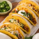 A row of five tacos filled with meat, cheese, sour cream, and cilantro, served on a wooden board. A bowl of sliced jalapeños and a bowl of white sauce are in the background.