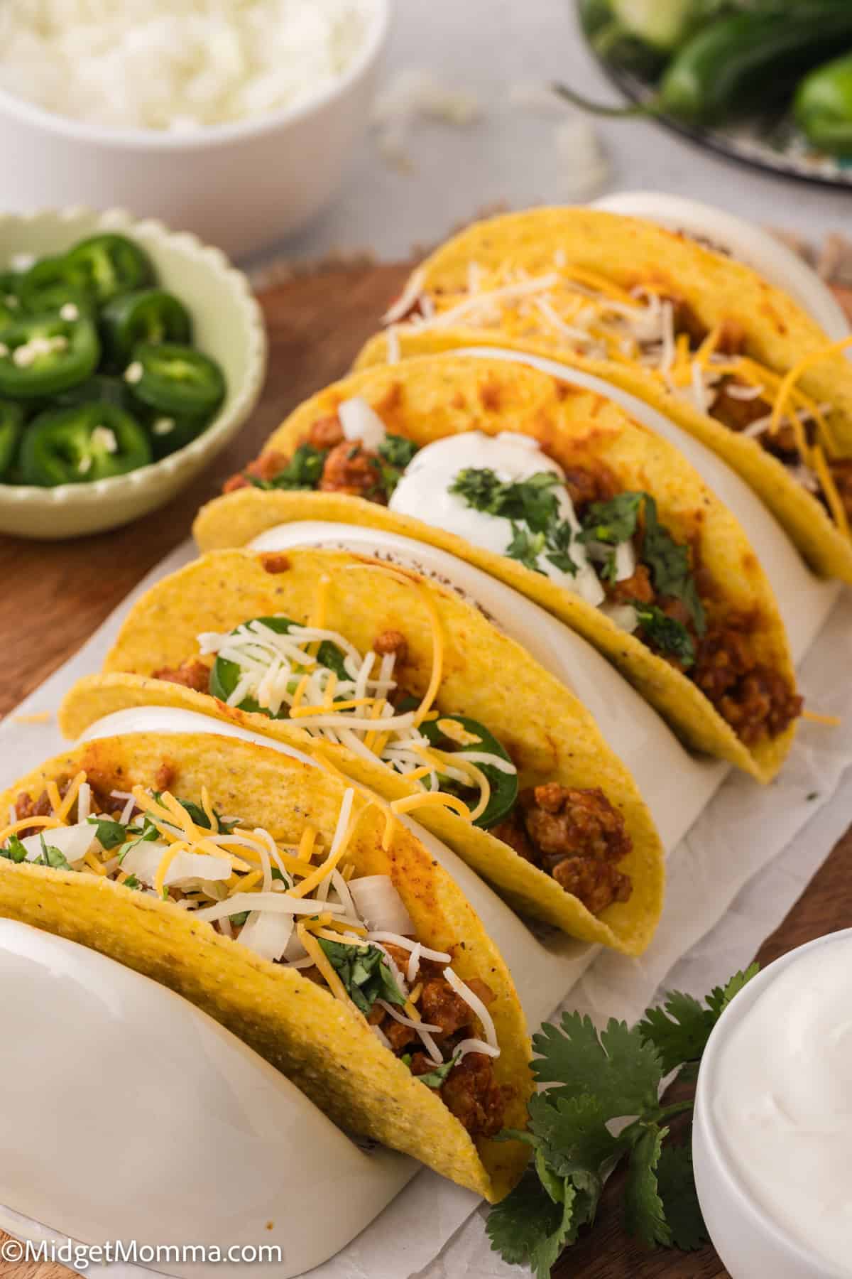 A row of five tacos filled with meat, cheese, sour cream, and cilantro, served on a wooden board. A bowl of sliced jalapeños and a bowl of white sauce are in the background.