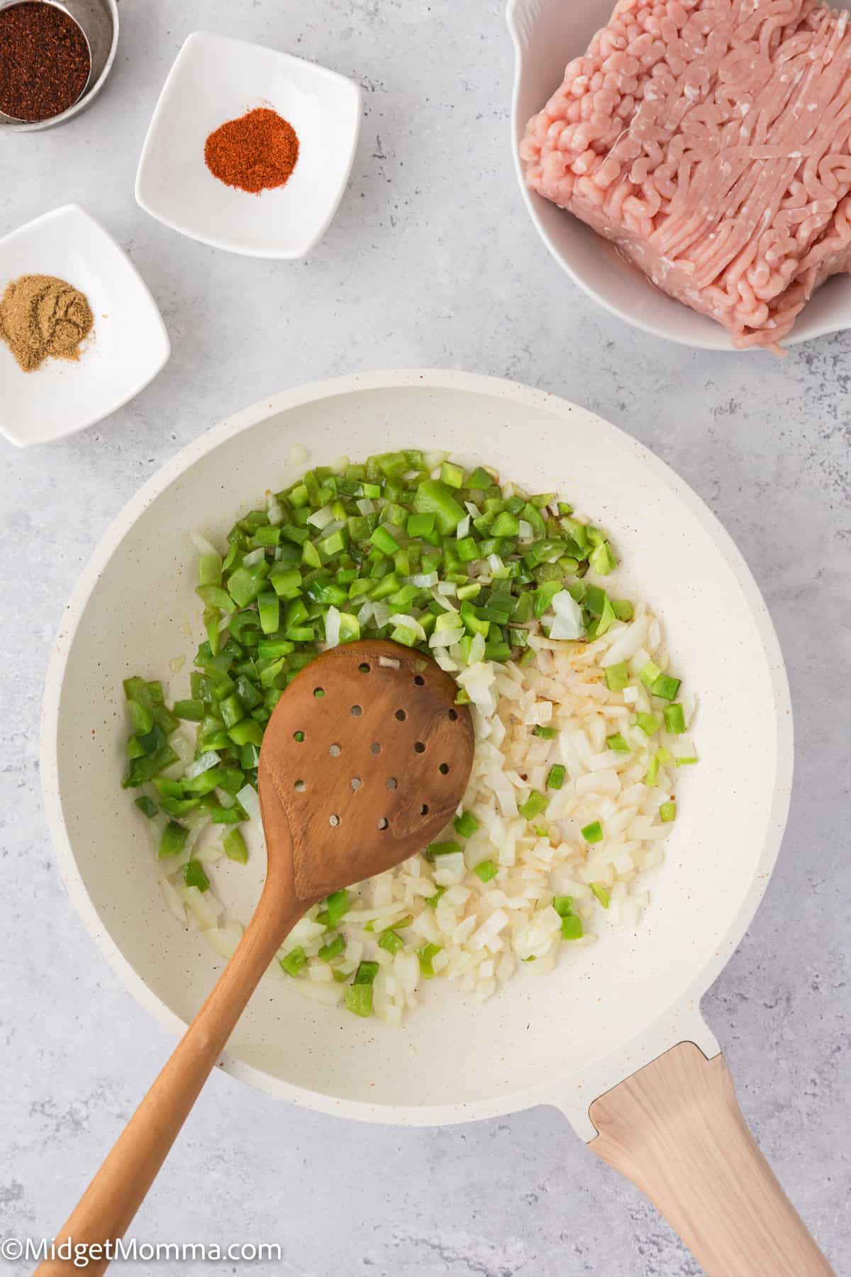 Chopped onions and green peppers cooking in a pan with a wooden spoon. Ground meat and spices are placed in small bowls nearby.