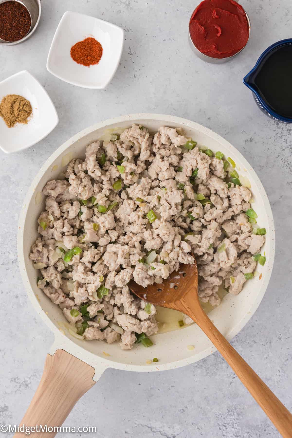 Ground chicken and chopped green peppers cooking in a white skillet, surrounded by bowls of spices and a small pitcher on a gray surface.