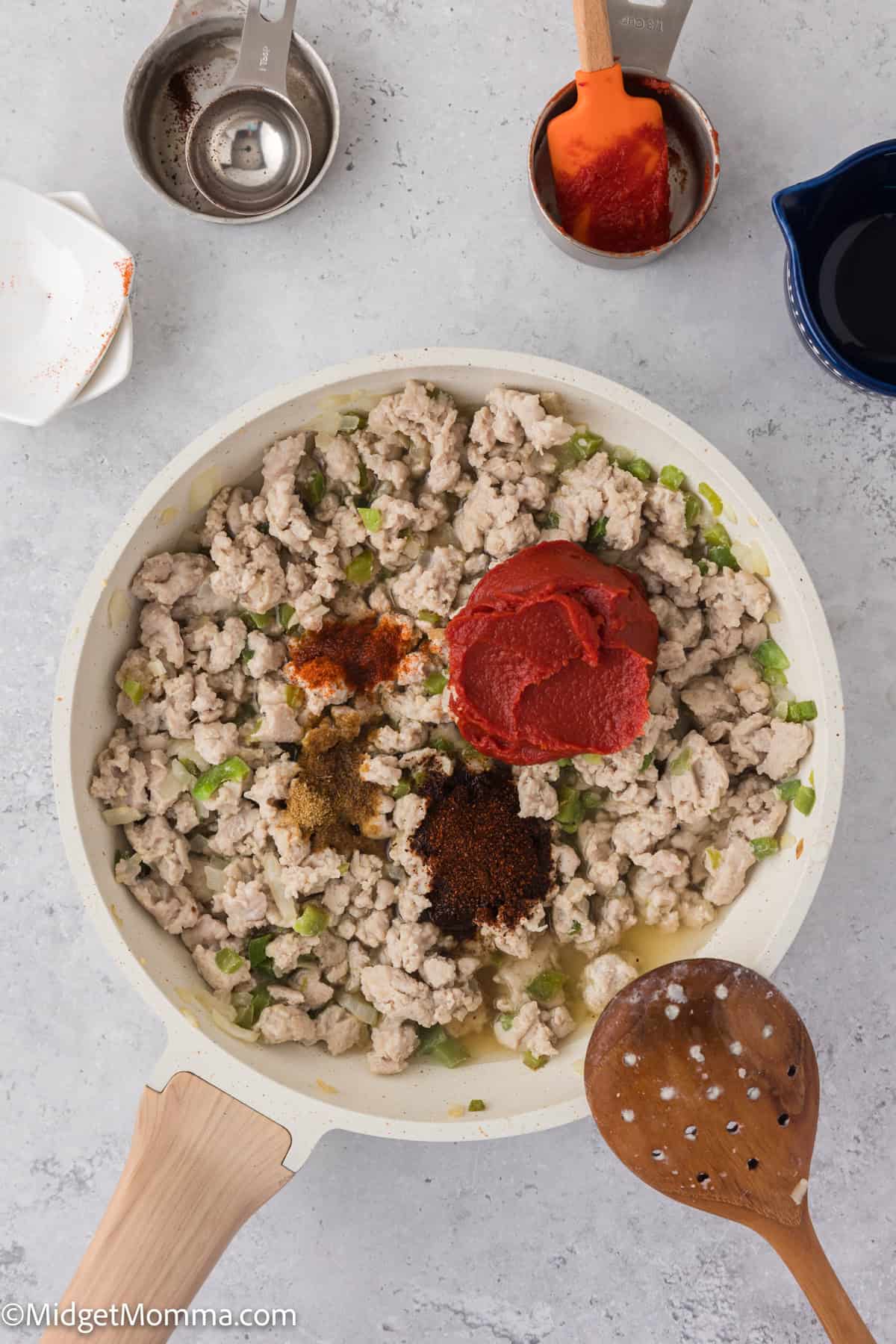 A skillet with cooked ground turkey, tomato paste, and various seasonings on a countertop. Nearby are measuring cups and a wooden spoon.