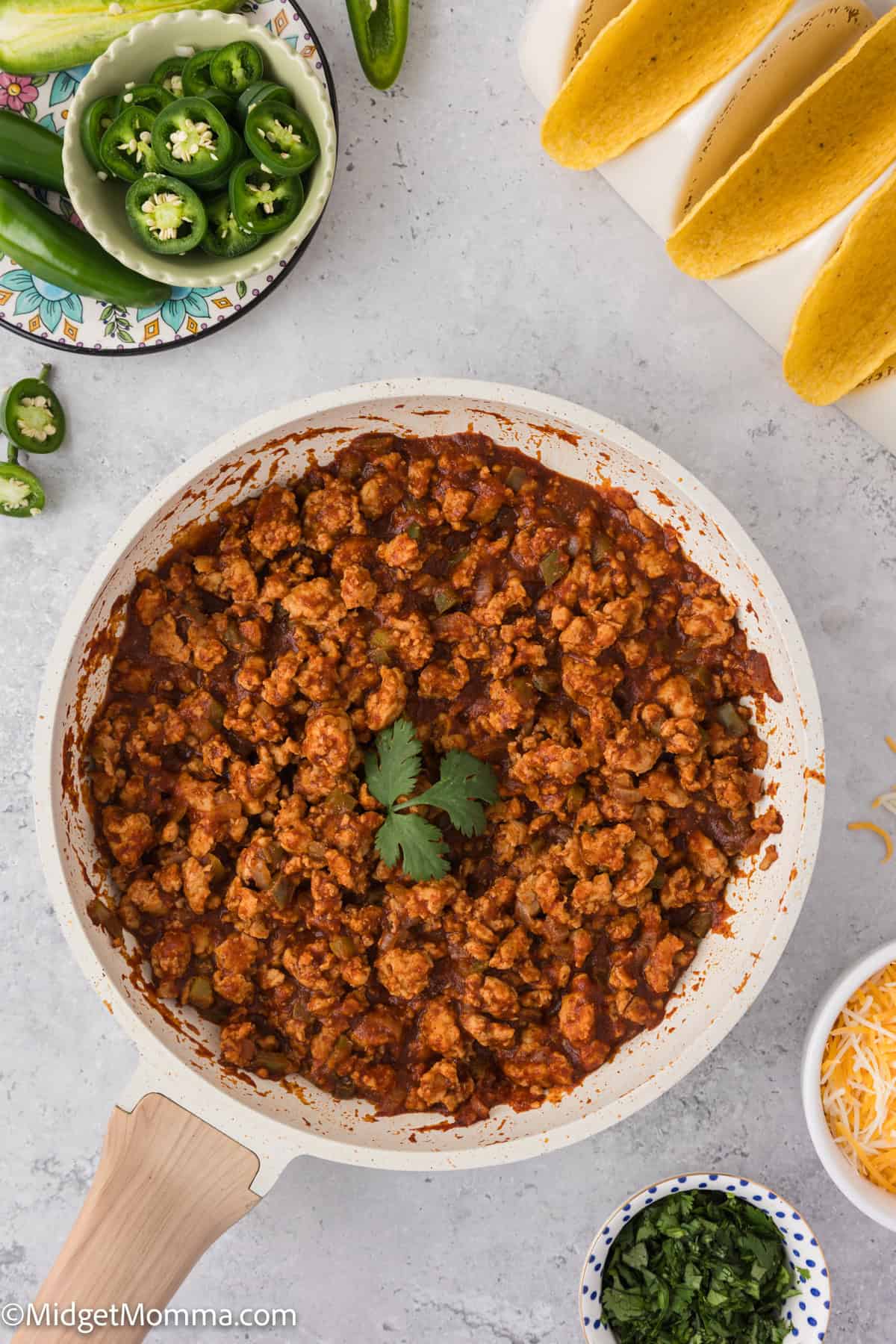 A skillet filled with cooked ground chicken taco meat topped with cilantro, surrounded by sliced jalapeños, cheese, lettuce, and taco shells on a light countertop.