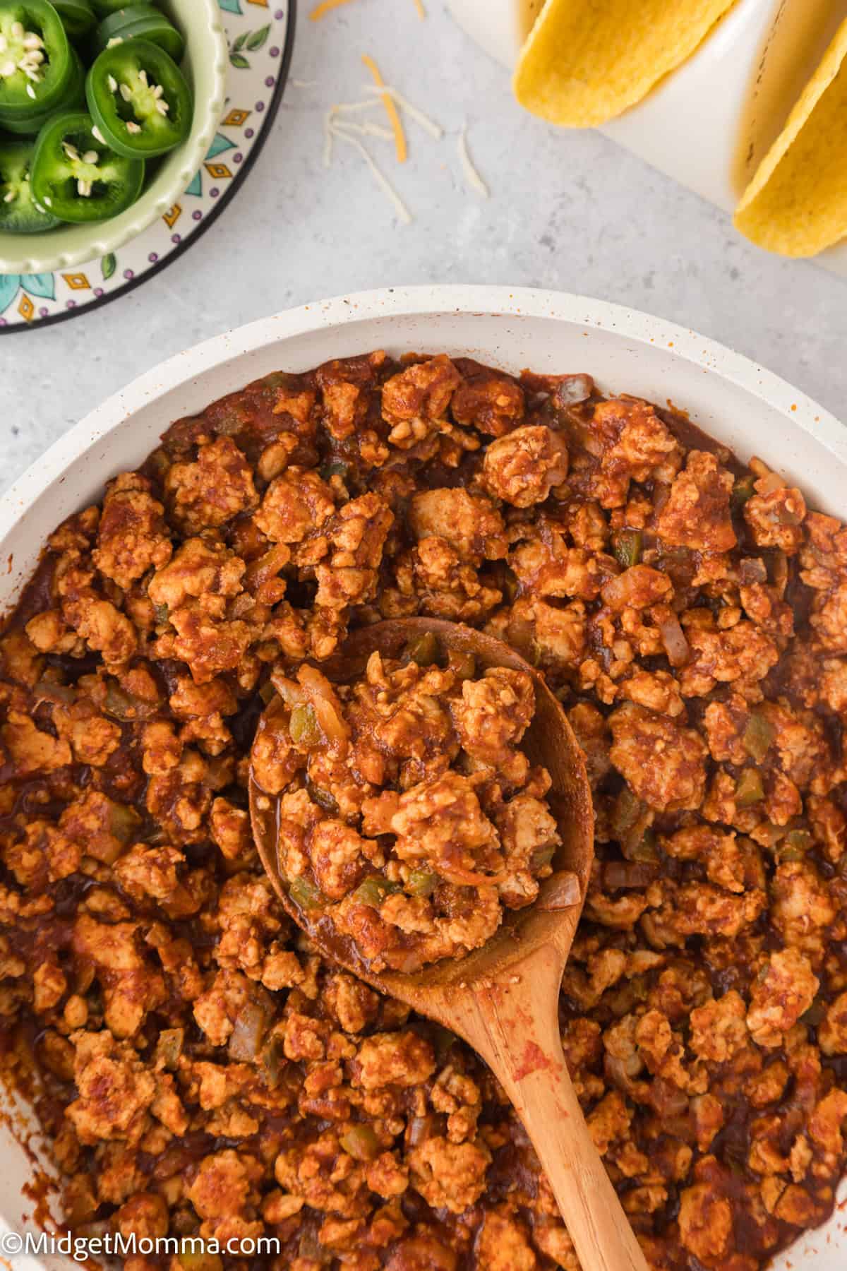 Pan of cooked taco meat with a wooden spoon, sliced jalapeños in a dish, and taco shells on the side.