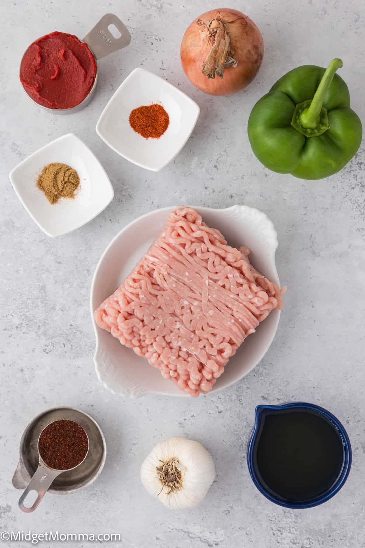 Ingredients on a table: ground turkey, green bell pepper, onion, garlic, spices in small dishes, tomato paste in a measuring cup, and a liquid in a blue cup.