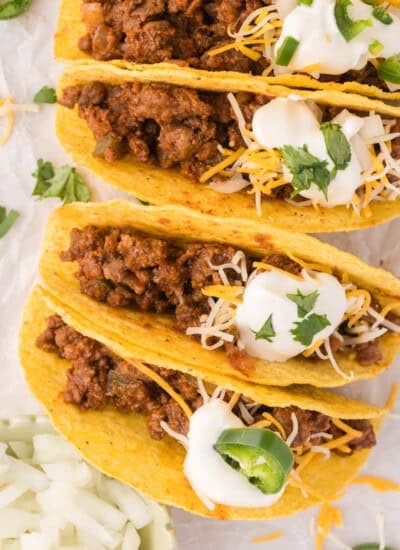 Three hard-shell tacos filled with seasoned ground meat, topped with shredded cheese, sour cream, and sliced jalapeños, garnished with cilantro.