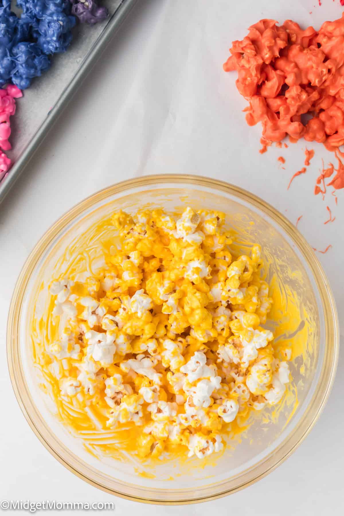 A glass bowl filled with yellow-colored popcorn sits next to a tray of blue and purple popcorn and a pile of red popcorn on a white surface.