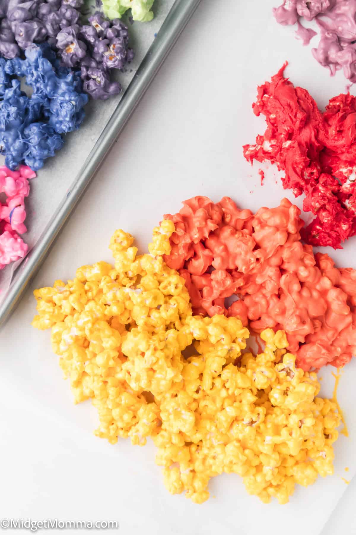 Colorful popcorn clusters in yellow, orange, red, pink, and purple are spread out on a surface, with some grouped on a baking sheet.