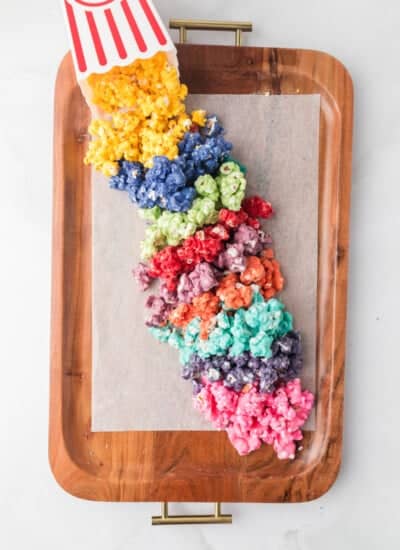 A tray with rainbow-colored popcorn spilling from a striped popcorn box onto a parchment-covered tray.