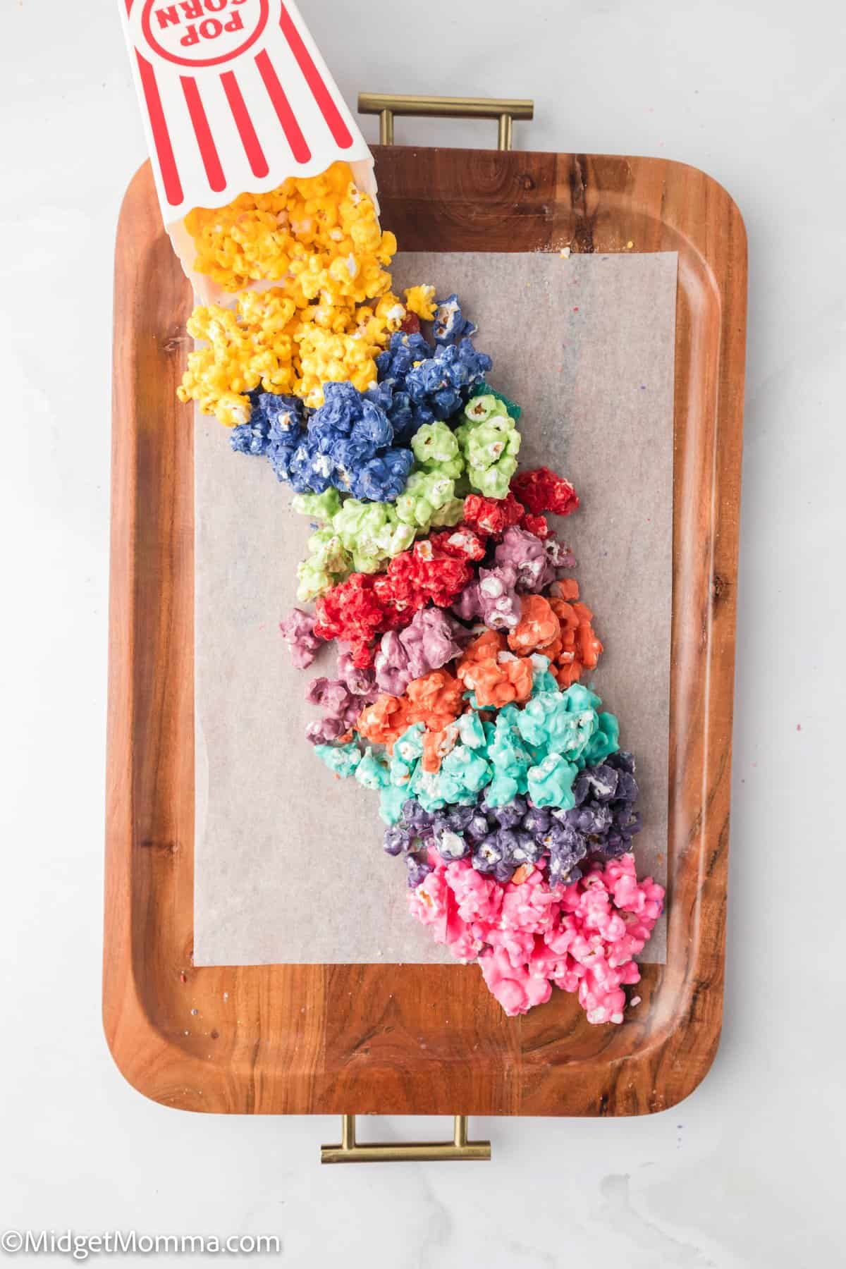 A tray with rainbow-colored popcorn spilling from a striped popcorn box onto a parchment-covered tray.