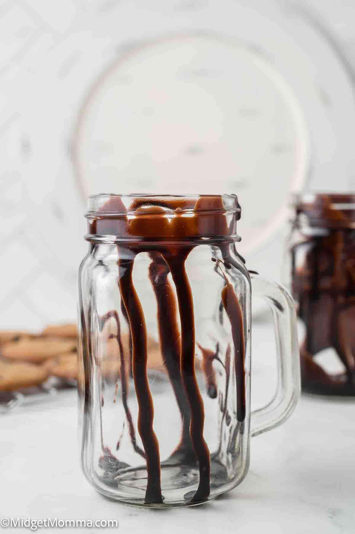A clear glass jar with a handle is drizzled inside with chocolate syrup, sitting on a white surface with blurred background items.