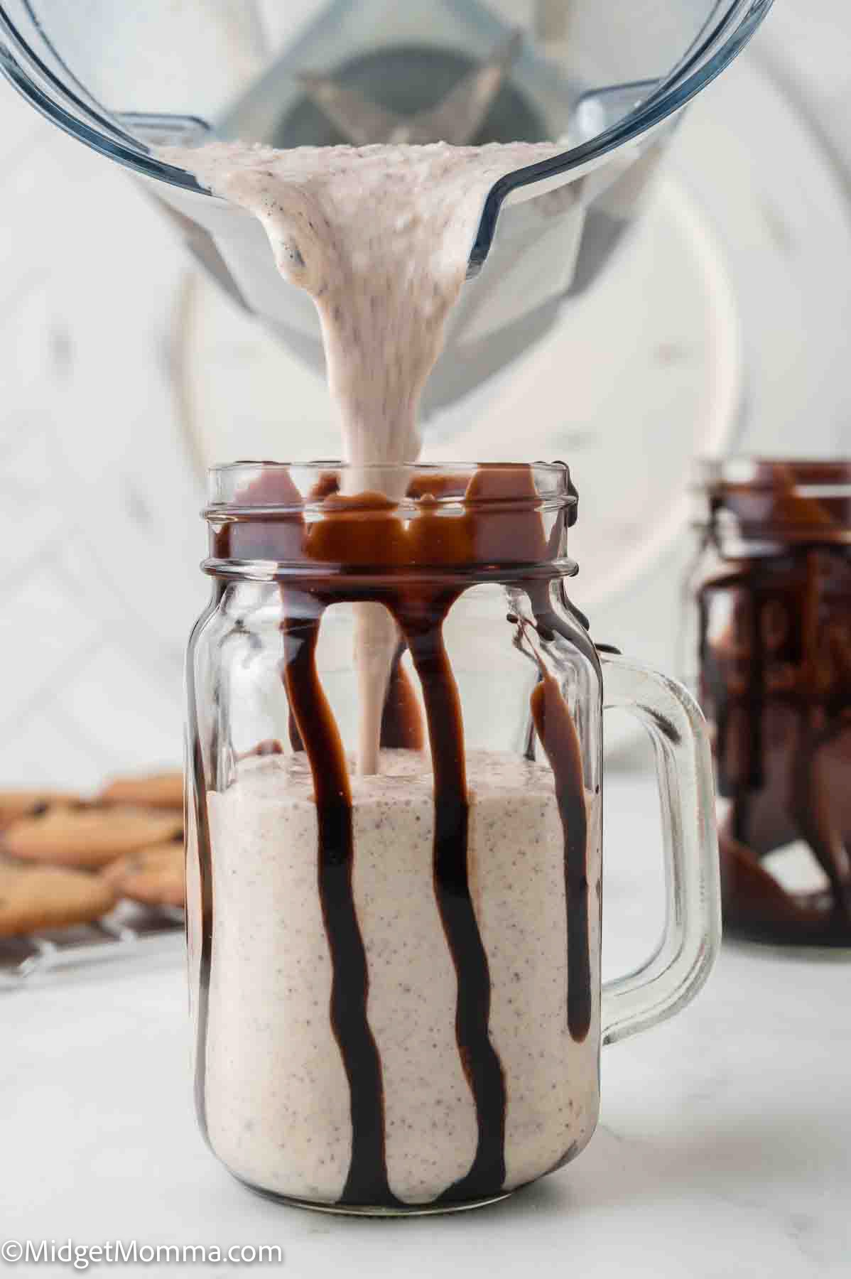 A thick milkshake is being poured from a blender into a mason jar decorated with chocolate syrup drizzles on the inside.