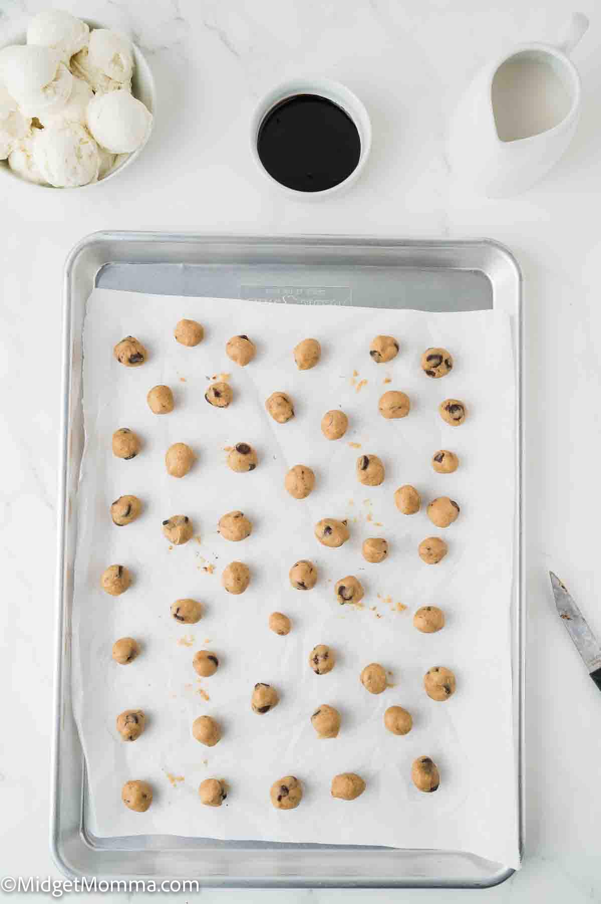 Baking sheet lined with parchment paper holding evenly spaced balls of chocolate chip cookie dough, with bowls of ingredients nearby.