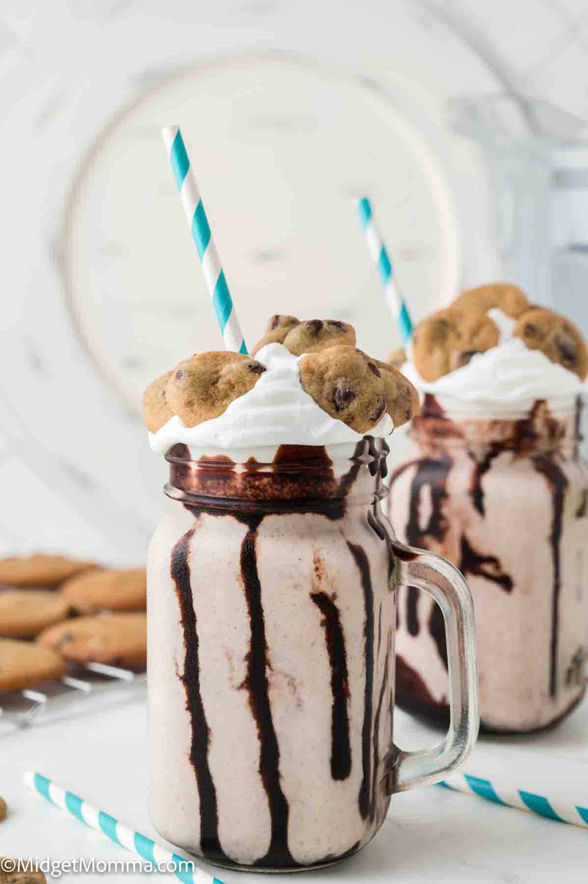 A mason jar filled with a chocolate-drizzled milkshake, topped with whipped cream and a chocolate chip cookie, served with a blue-striped straw.