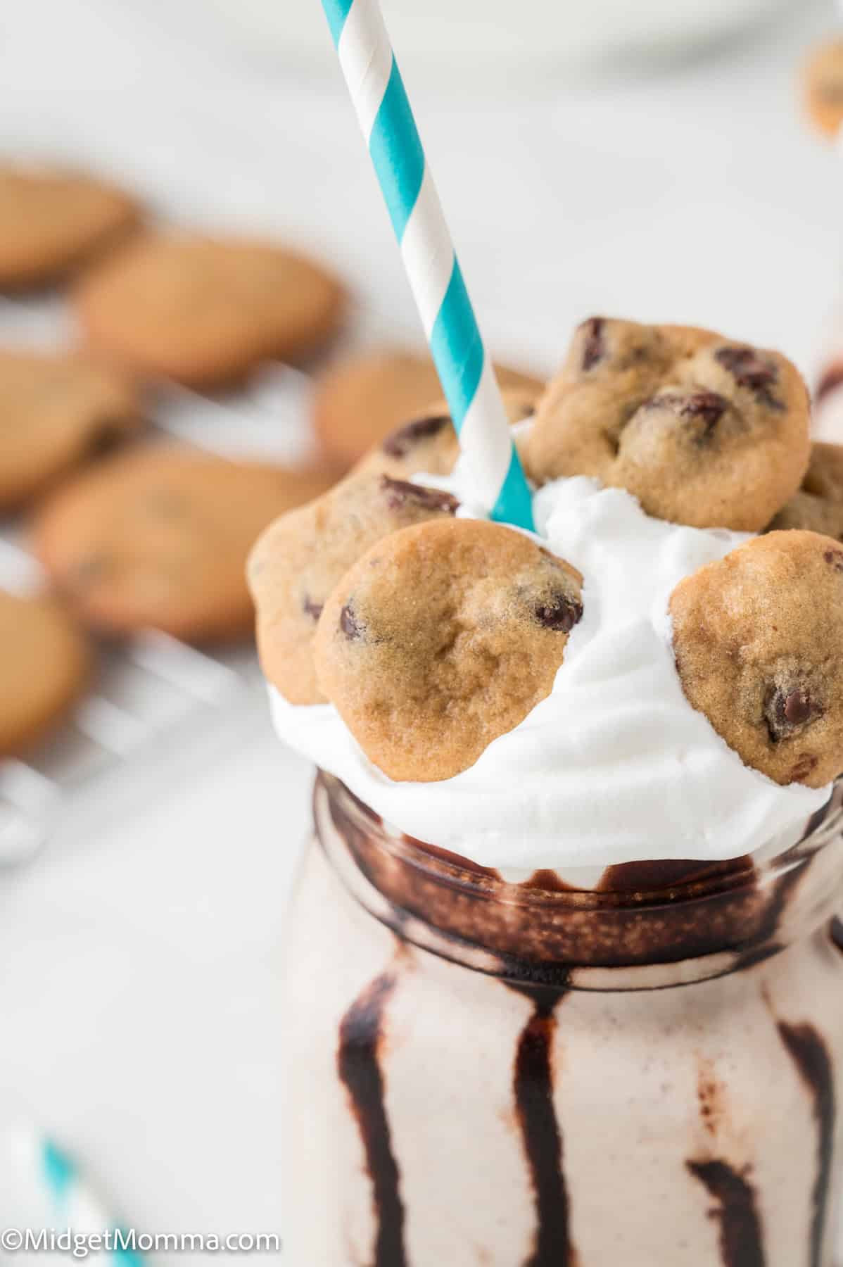A milkshake topped with whipped cream, mini chocolate chip cookies, and a blue striped straw, with chocolate drizzle inside the glass.