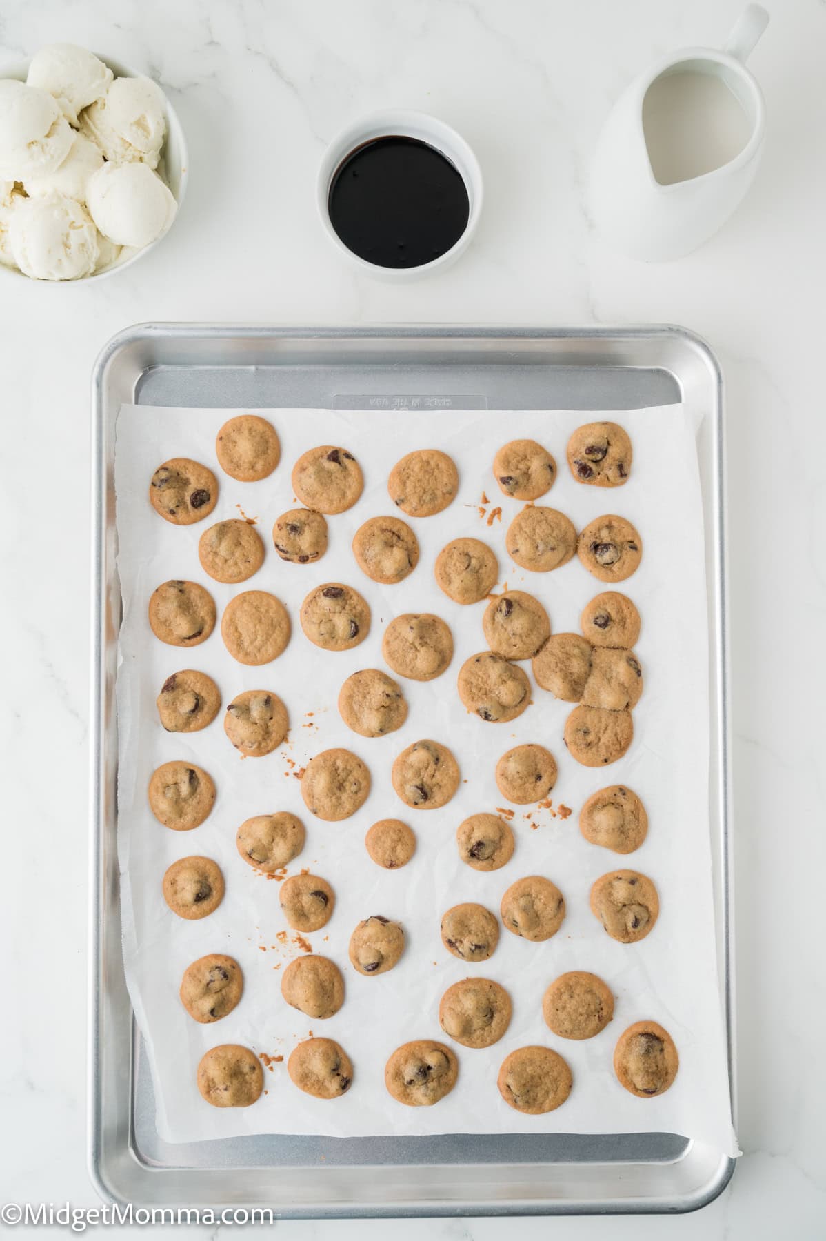 A baking tray with many small chocolate chip cookies on parchment paper, next to bowls of vanilla ice cream, chocolate sauce, and a pitcher of milk.