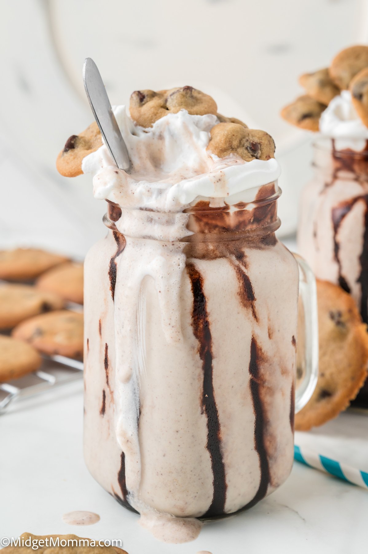 A mason jar filled with a cookie milkshake topped with whipped cream, chocolate syrup, and chocolate chip cookies, with a spoon sticking out.