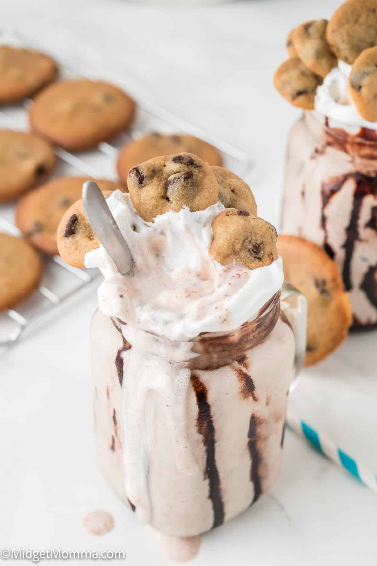 A glass jar filled with a chocolate milkshake, topped with whipped cream, chocolate chip cookies, and a spoon. More cookies are on a cooling rack in the background.