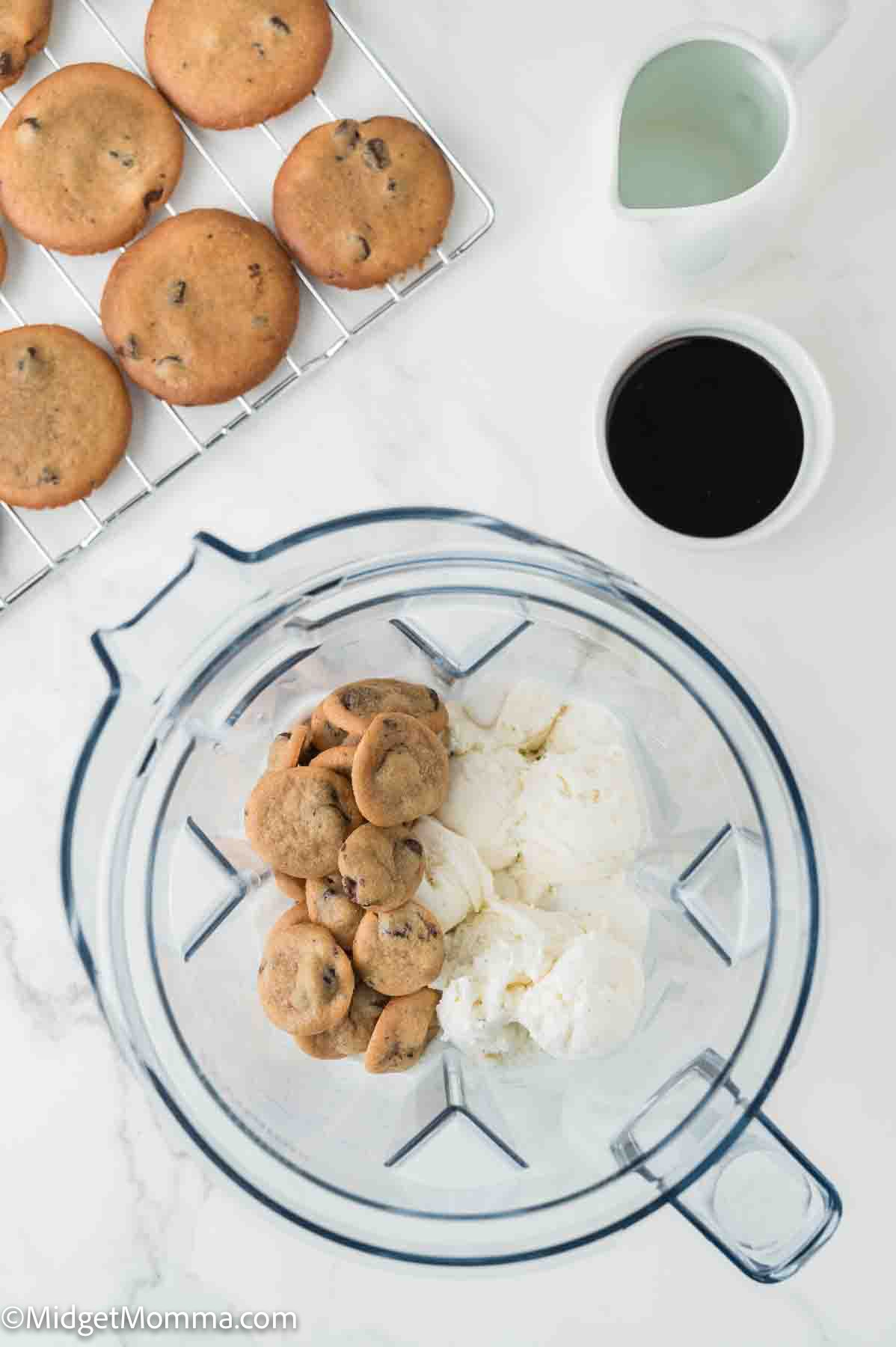 Chocolate chip cookies and vanilla ice cream in a blender, with more cookies on a cooling rack and cups of milk and coffee nearby on a white surface.
